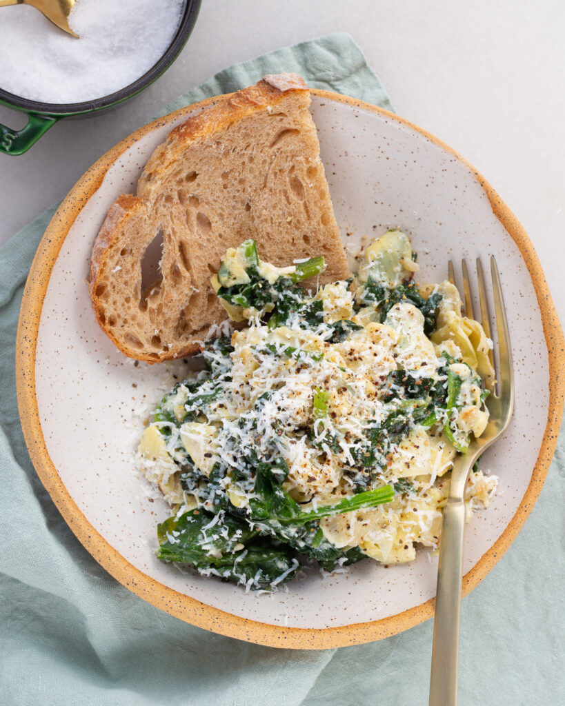 Spinach Artichoke Bowl plated for one, with a slice of bread and a fork. A salt cellar with a gold spoon is just partially out of frame.